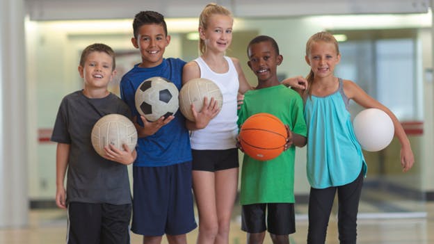 group of five boys and girls in a gym holding balls for various sports